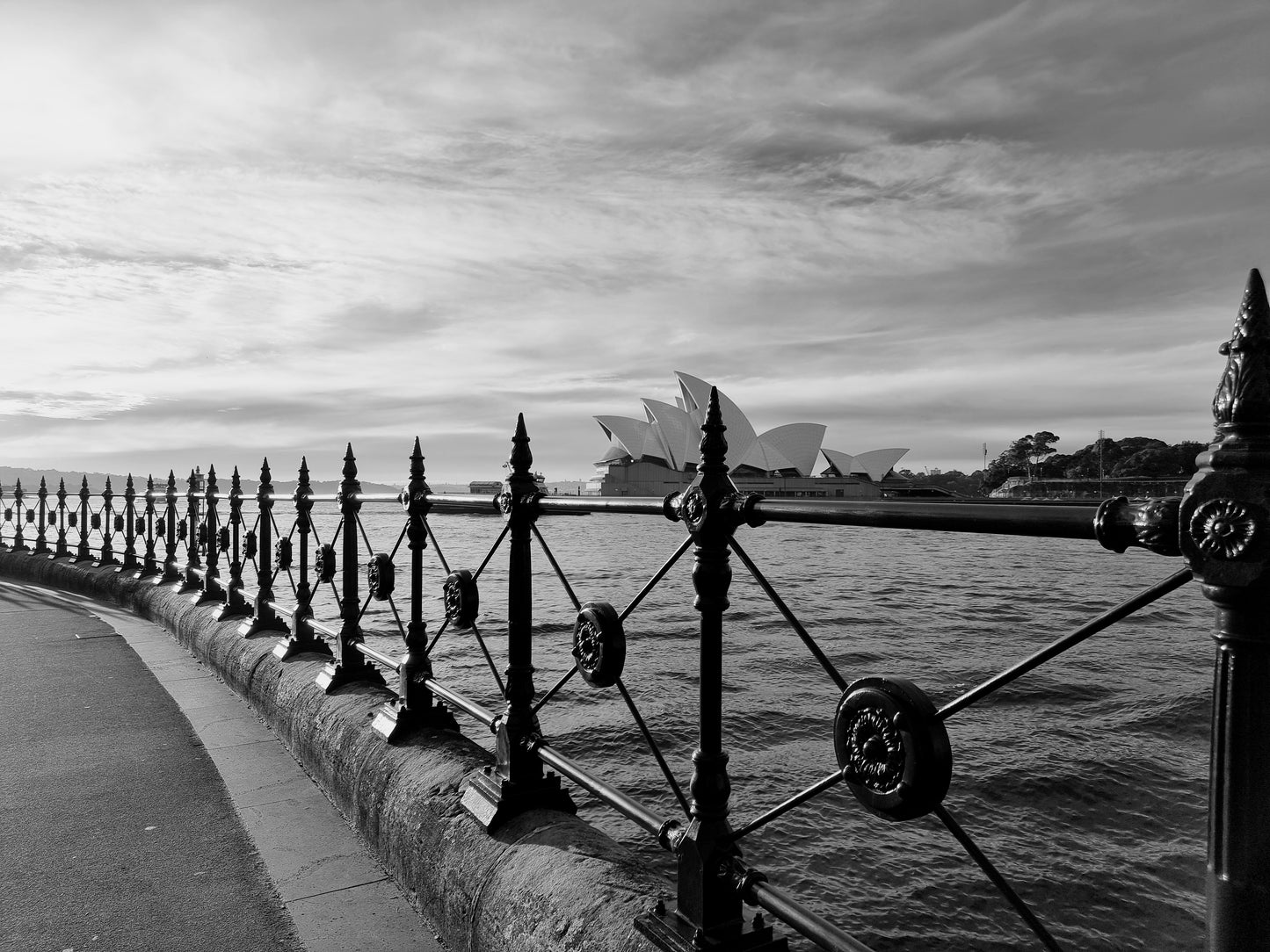 View form The Rocks through the ornate iron fencing, across Circular Quay, capturing the sail-like structure of the Sydney Opera House in black and white.