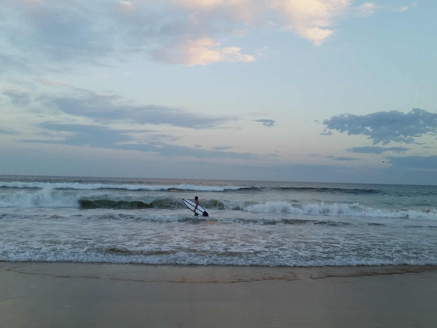 In the pale light of day a lone surfer in the surf at the shore line as the waves gently wash in.