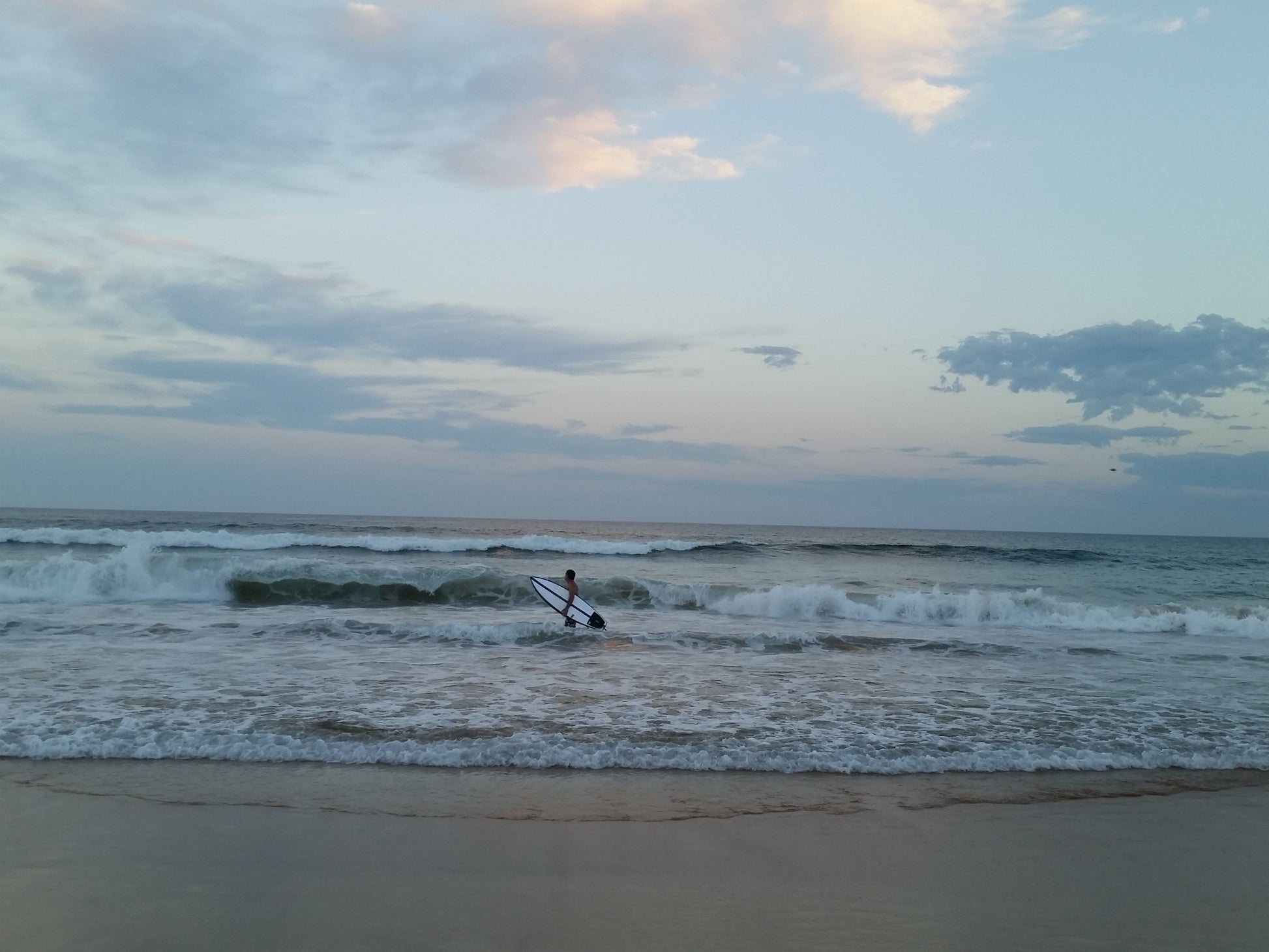 In the pale light of day a lone surfer in the surf at the shore line as the waves gently wash in. 