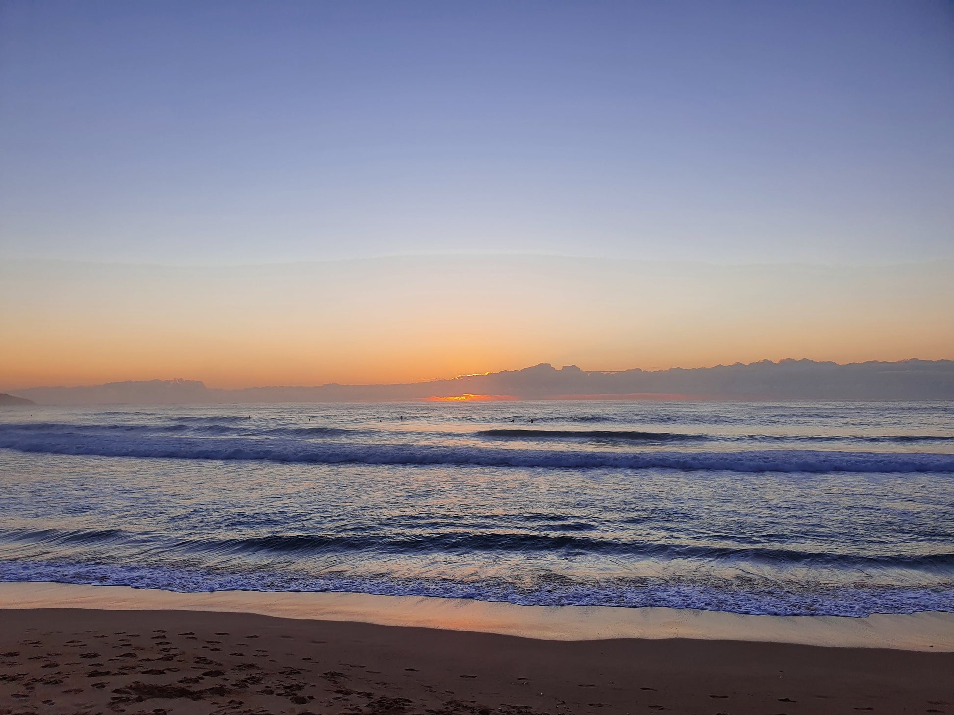 Pastel colours of dawn fill the sky and ocean, with white capped wave rolling to the shore.